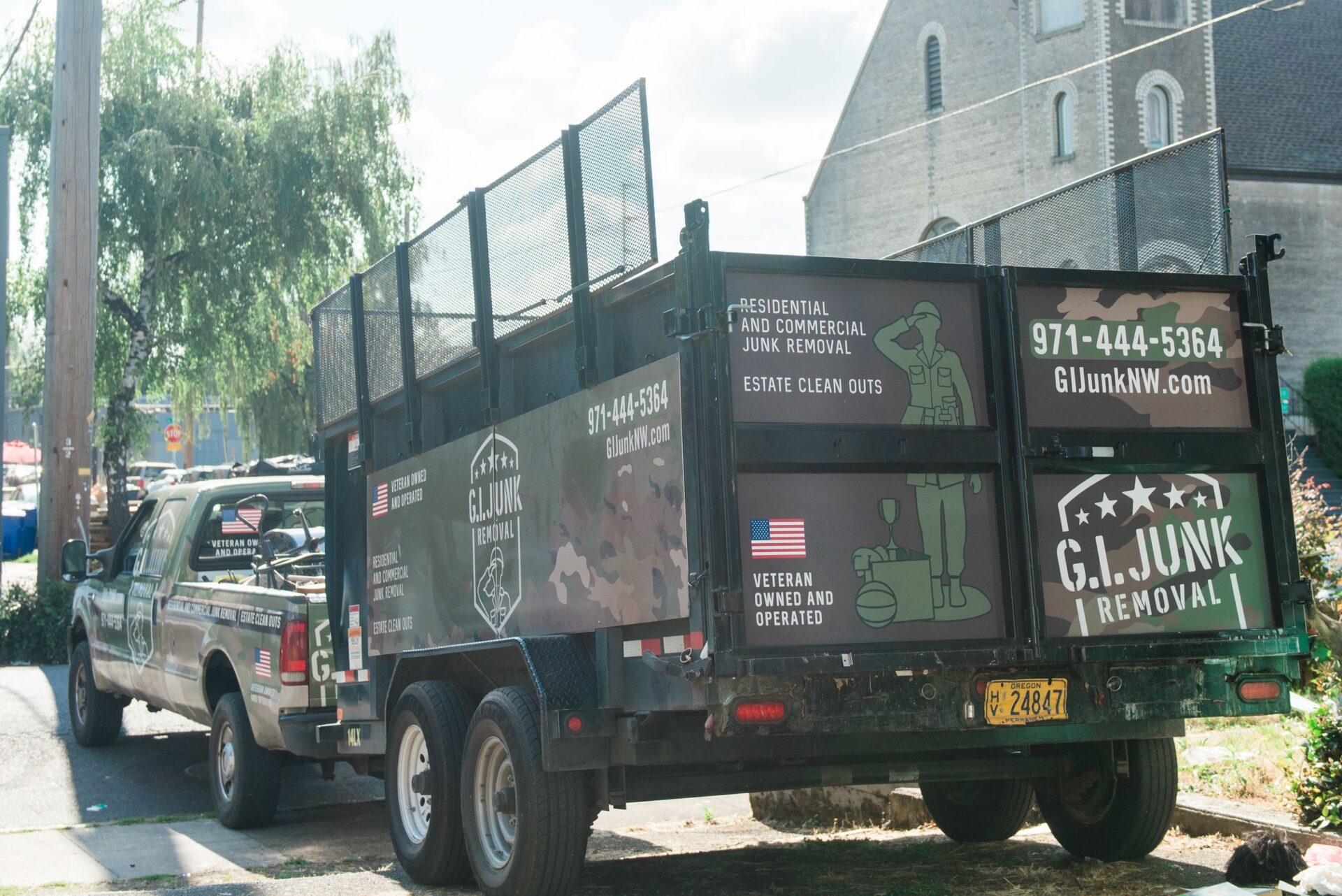 G.I. Junk Removal branded trailer and pickup truck parked on a street, showing camo-wrap graphics and contact info
