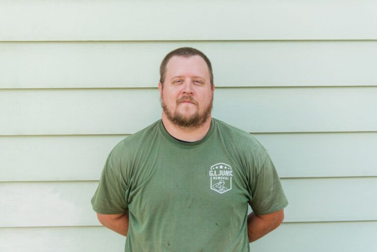 G.I. Junk Removal crew member in branded olive green t-shirt standing against light green siding