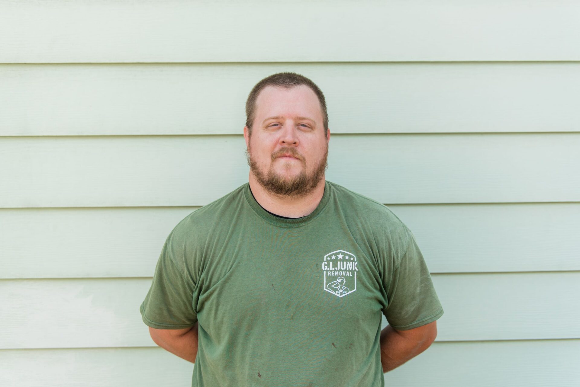 G.I. Junk Removal crew member in branded olive green t-shirt standing against light green siding